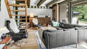 Un salon chaleureux avec une échelle en bois, une cheminée, un canapé en cuir et un coin repas offrant une vue sur l’extérieur.