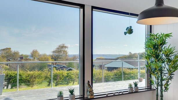 A room with large windows overlooking a garden and houses. There is a hanging light fixture and several potted plants on the windowsill. - "Clarita" - 300m to the fjord by Interhome (Roslev)