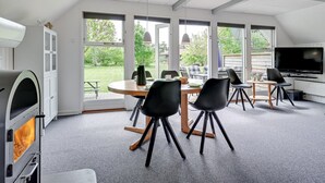 Eine moderne Küche mit Backofen, ein Essbereich mit rundem Tisch und schwarzen Stühlen, große Fenster mit Blick auf den Garten und ein flachbildfernseher an der Wand.