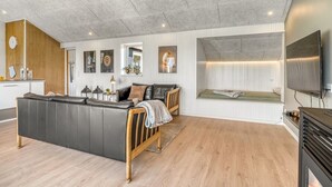 Een moderne woonruimte met een grote bank, een houten bankje, een inbouwbank met uitzicht, een flatscreen-tv en een keuken met een houten wand en een klok.