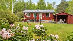 A red wooden house with a covered porch, a shed, and a swing set in a lush green garden.