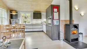 A modern kitchen with a wood-burning stove, a dining area with a table and chairs, and a view of the outdoors through large windows.
