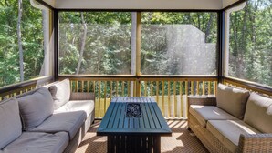 A covered outdoor seating area with wicker furniture and a central table, surrounded by large windows offering a view of a forested area. - Charming Pendleton Retreat ~ 5 Mi to Clemson! (Pendleton)