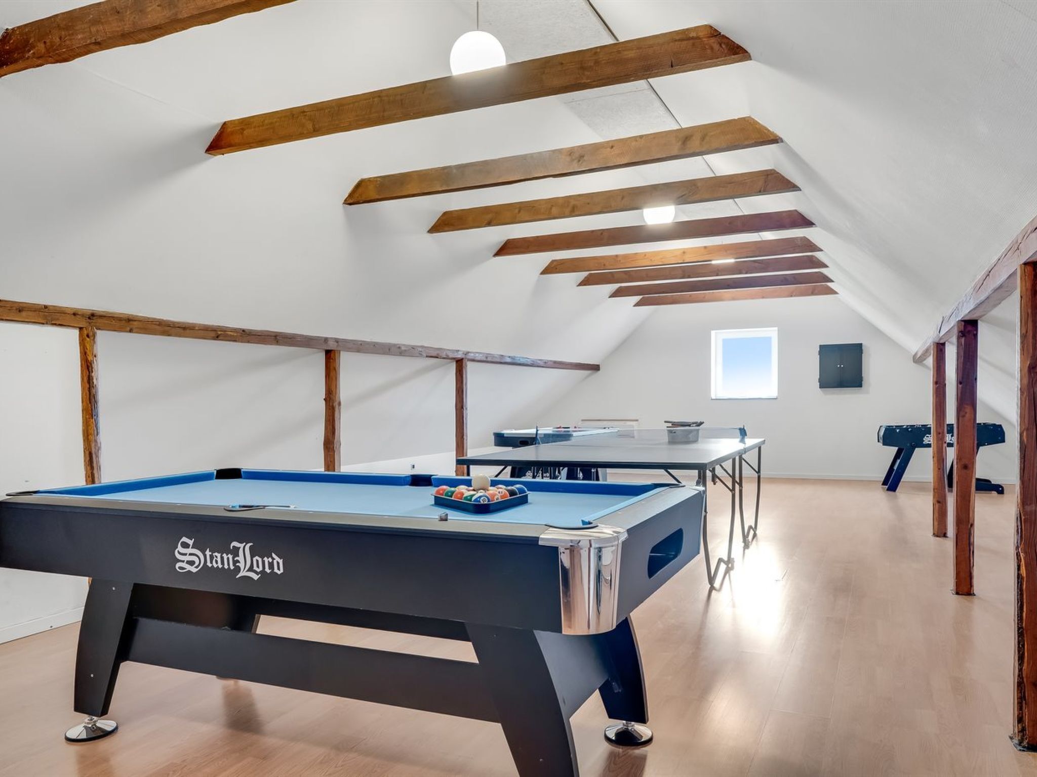 A pool table with a pool cue and balls, wooden beams on the ceiling, a table tennis table, and a window.