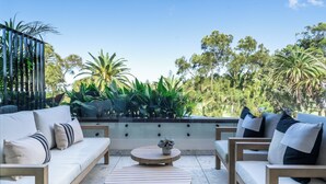 Un espacio al aire libre con sillón, sillas y una mesa redonda, rodeado de follaje y árboles.