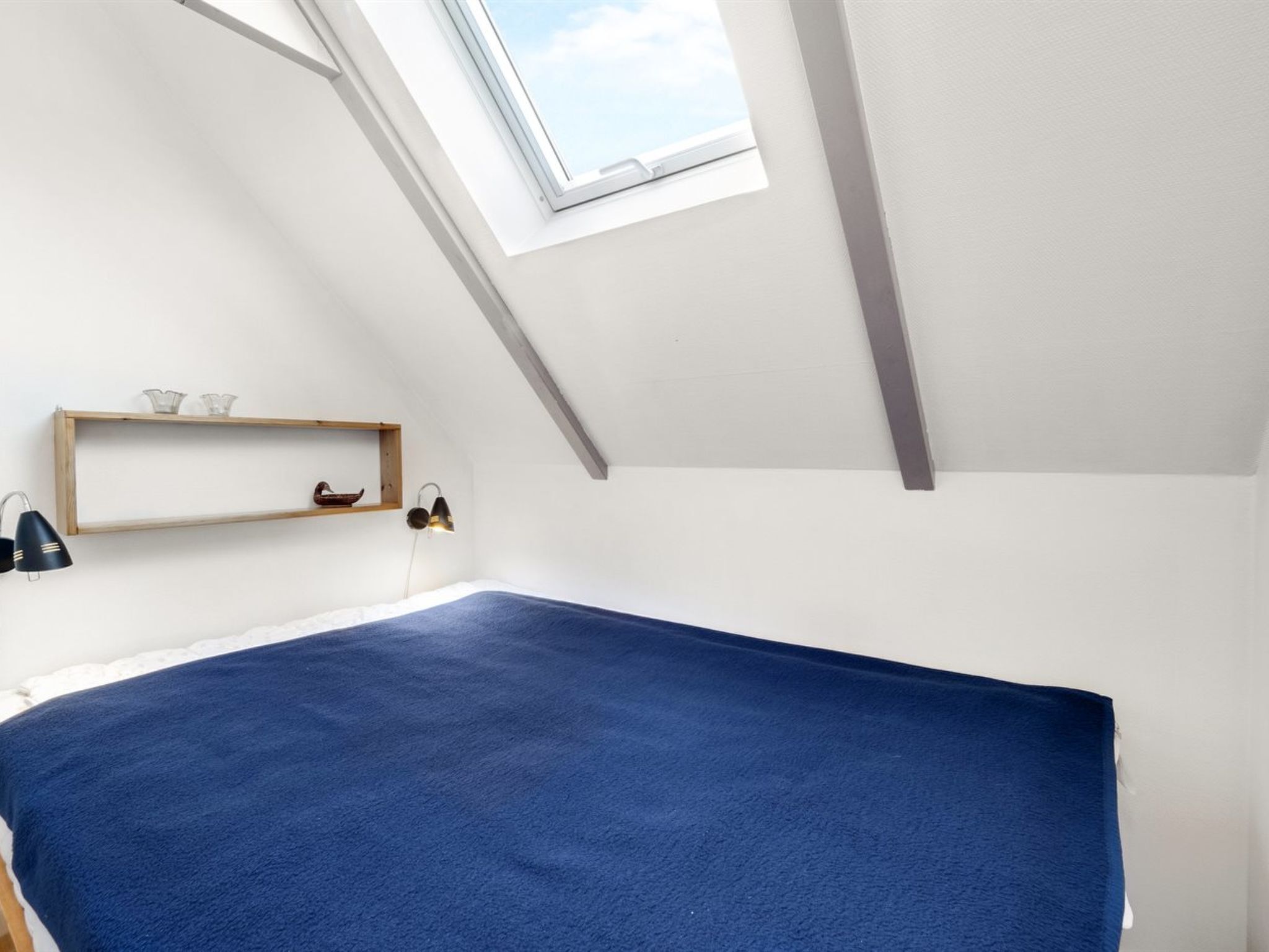 Une petite pièce blanche avec un plafond en pente et un puits de lumière. On y trouve une étagère en bois ornée d’objets décoratifs et une lampe fixée au mur.