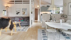 A modern interior with a wooden floor, a grey sofa, a dining table with chairs, and a kitchen area with cabinets and a hanging light fixture.