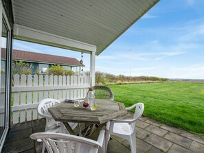 Eine überdachte Terrasse mit einem Holztisch und weißen Plastikstühlen, mit Blick auf ein grünes Feld und ein entferntes Gebäude.