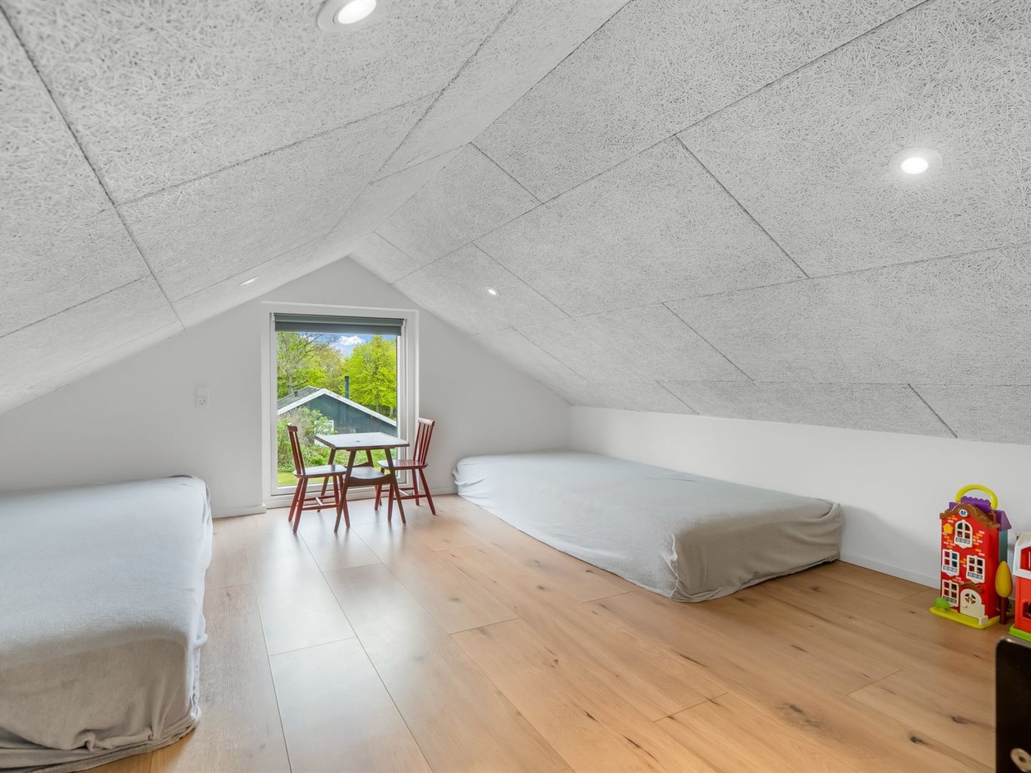 Ein kleines Zimmer im Dachgeschoss mit geneigter Decke, einem Fenster mit Blick ins Grüne, einem Tisch mit Stühlen und zwei Betten.