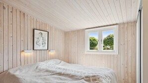 Quarto com paredes de madeira, cama com cobertor branco, quadro na parede e janela com vista para árvores.