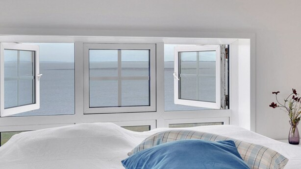 Een slaapkamer met een groot raam met uitzicht op zee, een bed met wit- en blauw beddengoed, en een bloevas op het nachtkastje.