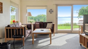 Ein Wohnzimmer mit einer Couch, einem Couchtisch und einer TV-Konsole. Ein großes Fenster bietet Blick auf Grünflächen und ein Gewässer.