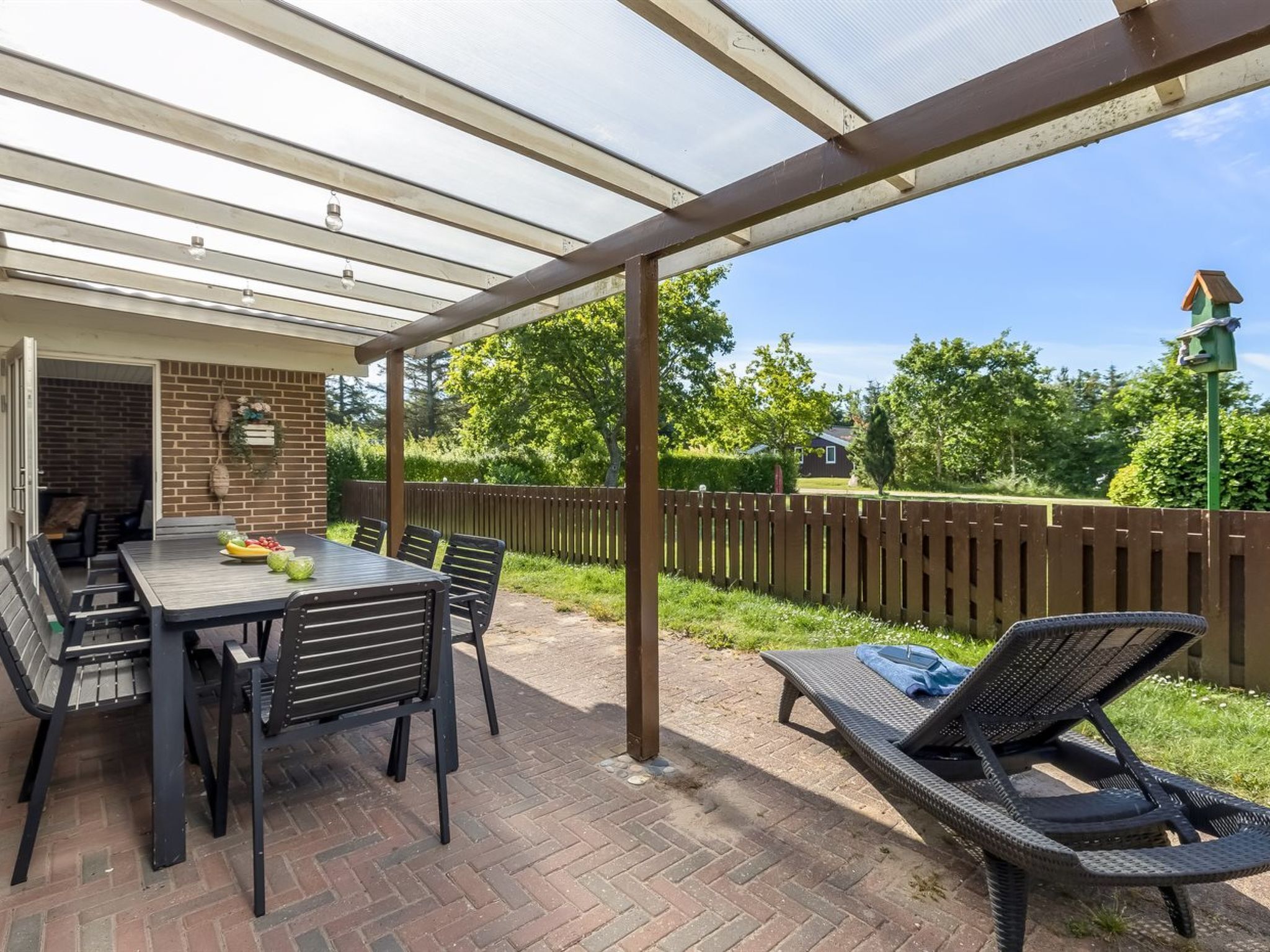 A covered patio with a table and chairs, a wicker lounge chair, and a wooden fence.
