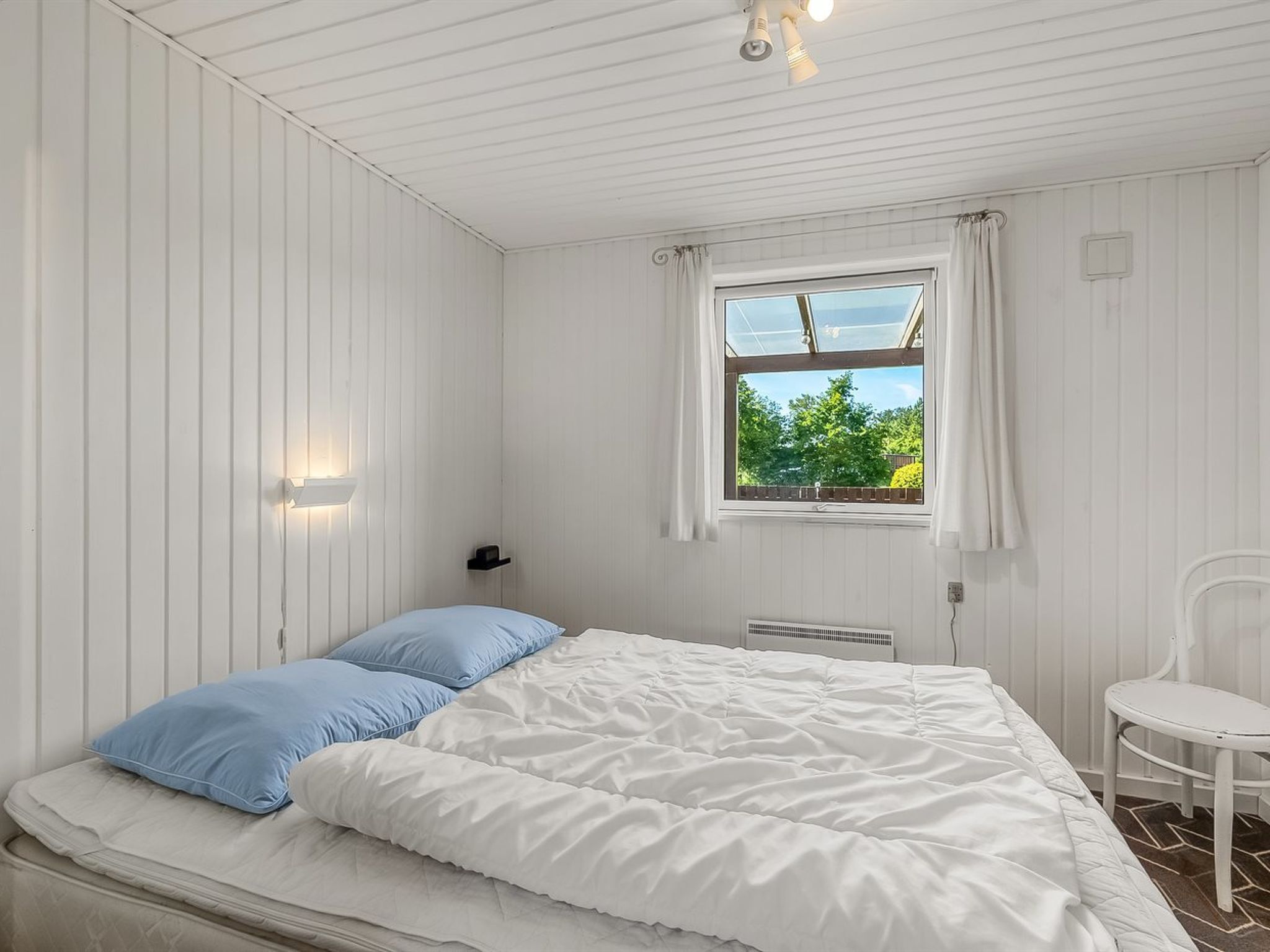 Un dormitorio con cama, ventana con cortinas, una silla y una lámpara fijada a la pared.