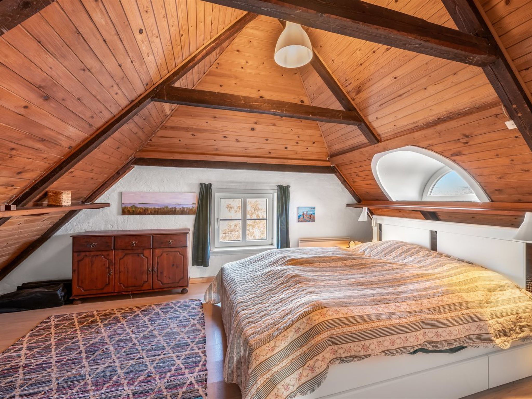 Une chambre confortable située sous les toits, avec un plafond en pente, des poutres apparentes, un lit recouvert d’une couvre-lit à motifs, une commode et une fenêtre offrant une belle vue.