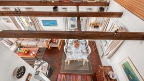 Amplia habitación tipo loft con techo alto, vigas de madera a la vista, zona de comedor con mesa y sillas, un rincón de descanso con sofá y vistas a la ciudad a través de amplios ventanales.