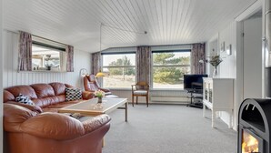 Ein gemütliches Wohnzimmer mit Ledersofa, Fernseher, Kamin und großen Fenstern mit Blick ins Freie.