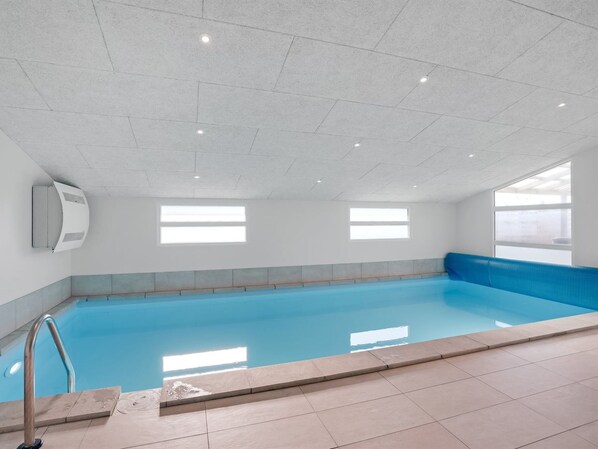 Una piscina interior con agua azul cristalina, suelo de baldosas y un banco a un lado.