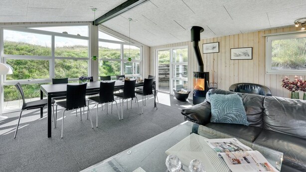 A modern living room with a fireplace, a dining area with chairs, and large windows offering a view of greenery. - "Jess" -  from the sea by Interhome (Ringkøbing)