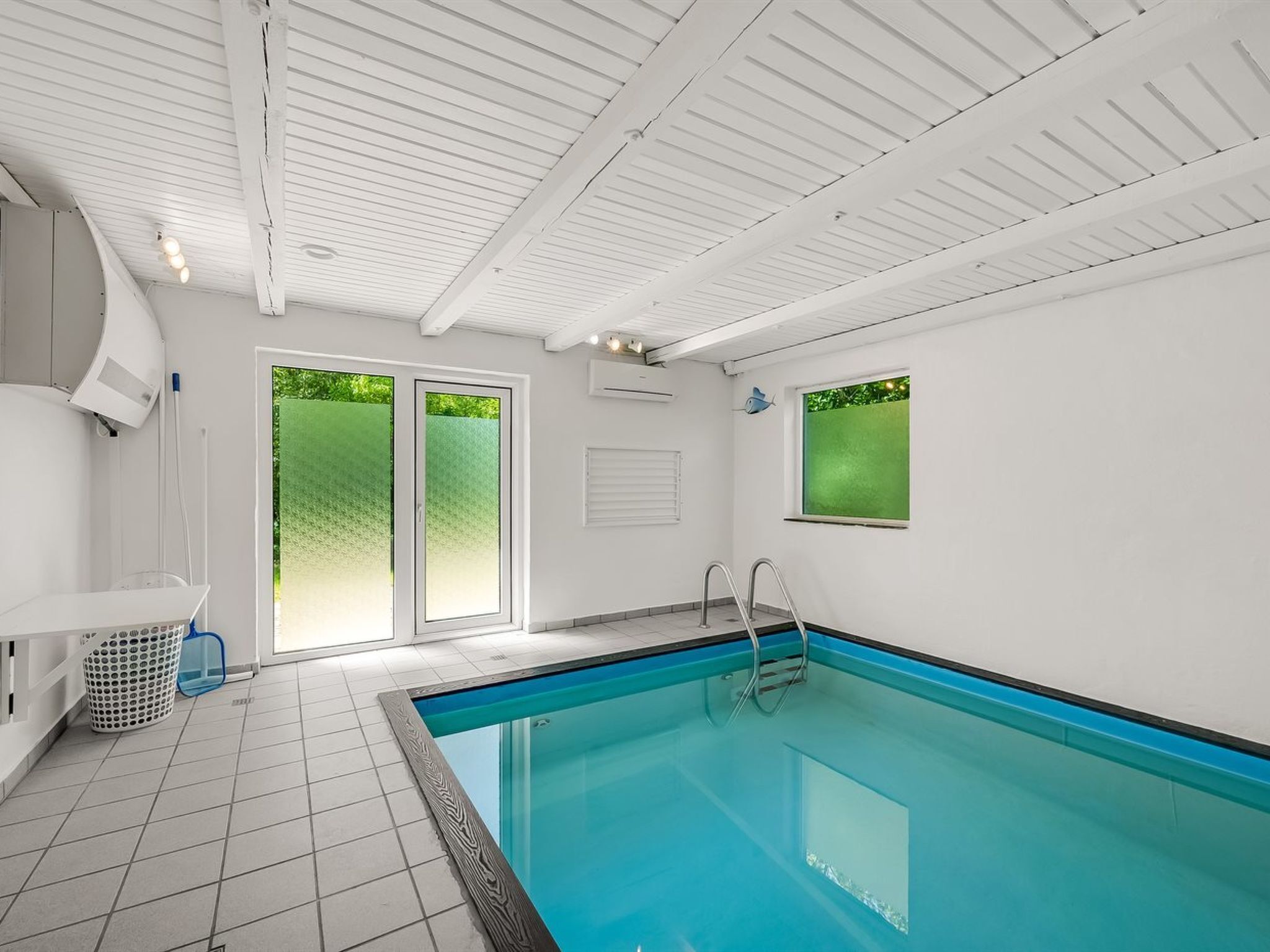Una piscina interior con suelo de baldosas, una escalera y una ventana con vistas a la vegetación.