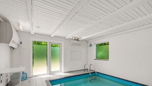 Une piscine intérieure avec un sol carrelé, une échelle et une fenêtre donnant sur un paysage verdoyant.