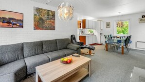 Un salon moderne avec un canapé gris, une table basse en bois sur laquelle on voit des fruits, un coin repas avec une table et des chaises, et une cuisine en arrière-plan.