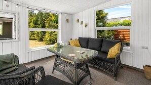 A sunlit room with a glass table, wicker chairs, a black sofa with yellow cushions, and large windows offering a view of greenery. - "Eira" - 350m to the fjord by Interhome (Tarm)