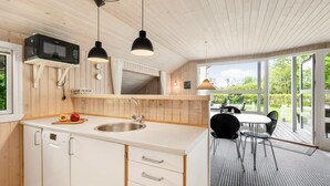 Une cuisine moderne avec des meubles en bois, un évier en acier inoxydable et un four à micro-ondes. On y trouve un coin repas avec une table et des chaises, et une vue sur l’extérieur grâce à de grandes fenêtres.