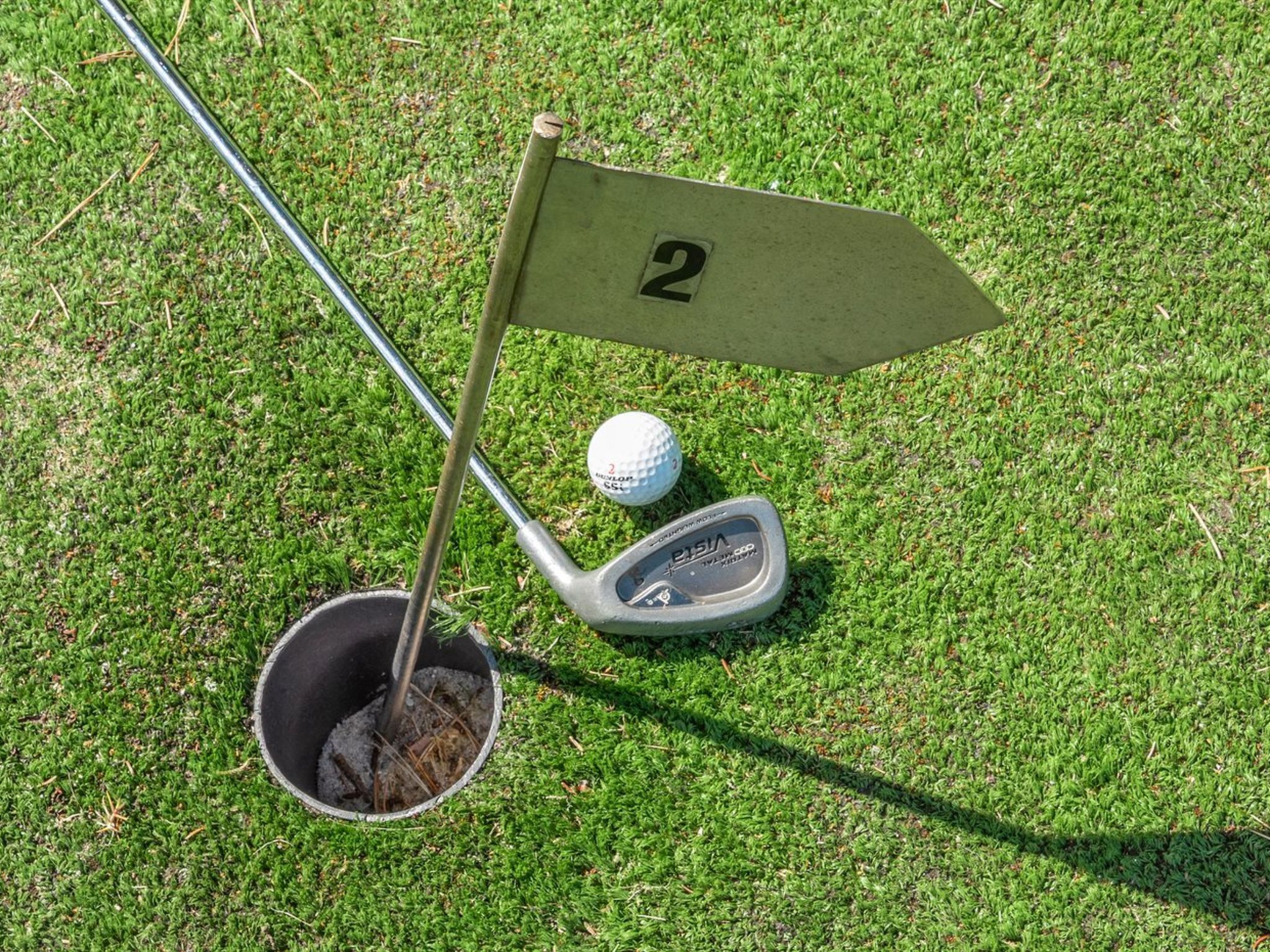 Ein Golfloch mit einer Fahne, die die Nummer 2 zeigt, einem Golfball und einem Putterschläger auf einer Rasenfläche.