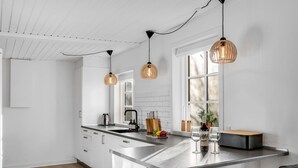 Cocina moderna con armarios blancos, una isla central, iluminación colgante y vistas al exterior gracias a grandes ventanas.
