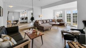 Amplia sala de estar con un sofá, sillones y una mesa de centro. Se ve una cocina al fondo y un ventanal que proporciona luz natural.