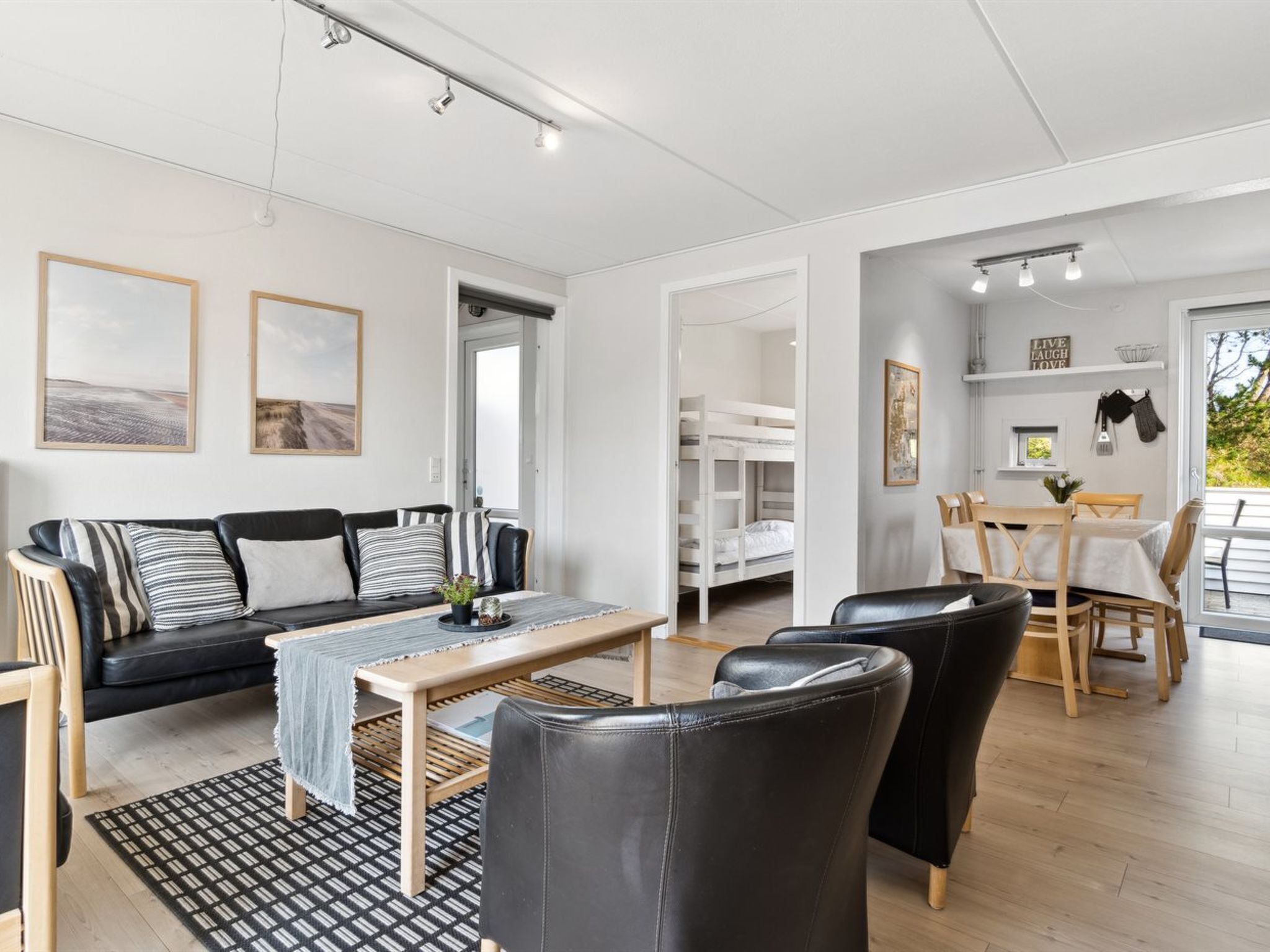 Un salon moderne avec un canapé, une table basse et des fauteuils, un coin repas avec une table et des chaises, et un lit superposé à l’arrière-plan.
