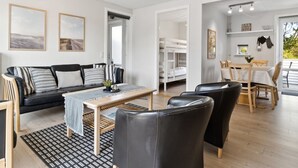 Un salon moderne avec un canapé, une table basse et des fauteuils, un coin repas avec une table et des chaises, et un lit superposé à l’arrière-plan.