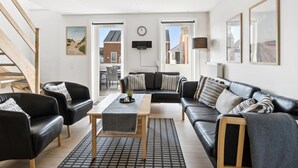 Un salon moderne avec un ensemble de canap és en cuir noir, une table basse en bois et un tapis à motifs. On aperçoit un escalier sur la gauche, une vue sur l’extérieur à travers une porte vitrée et une horloge au mur.