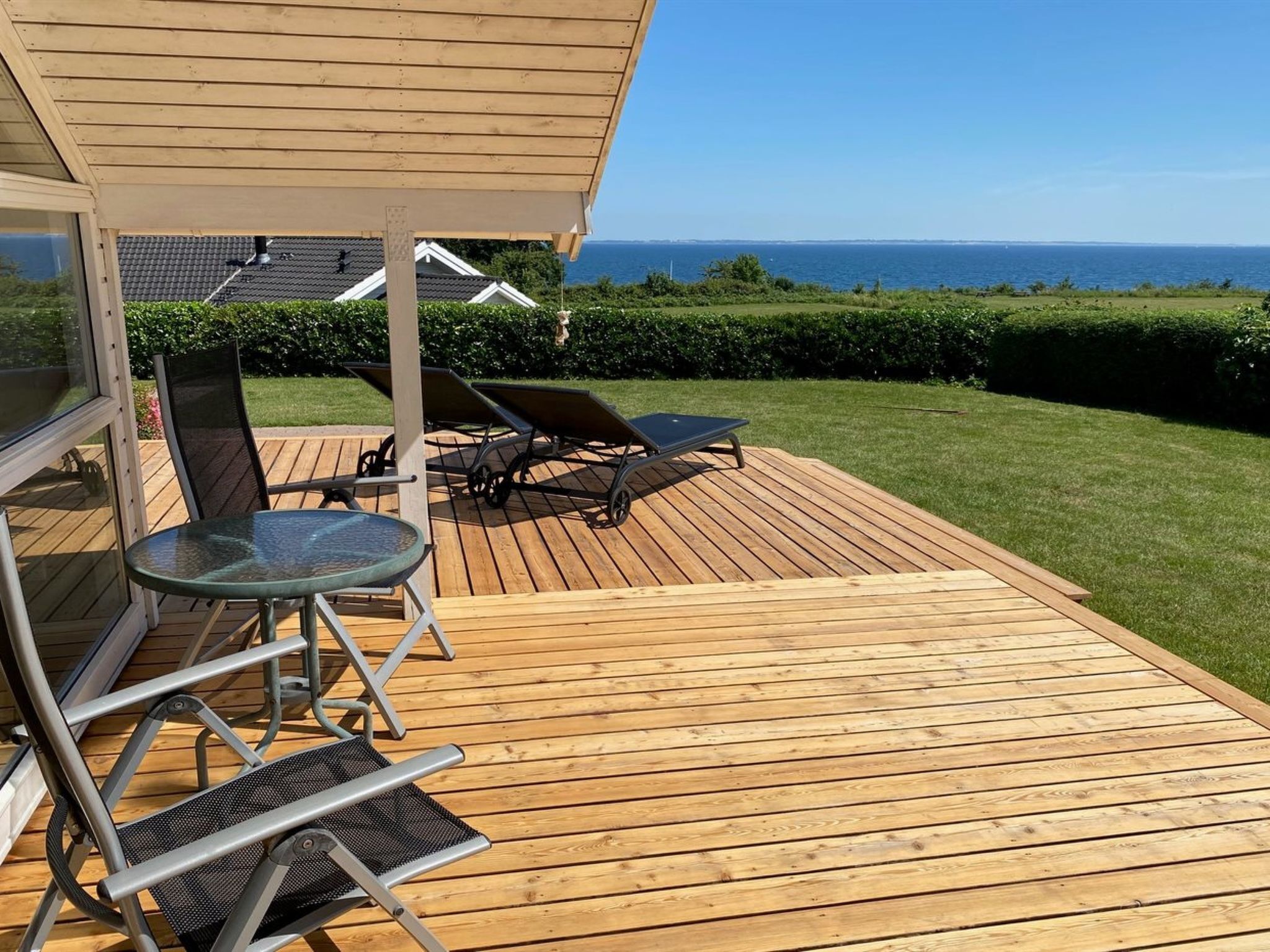 Eine Holzterrasse mit Glastür, einem Tisch, zwei Stühlen und zwei Liegestühlen.