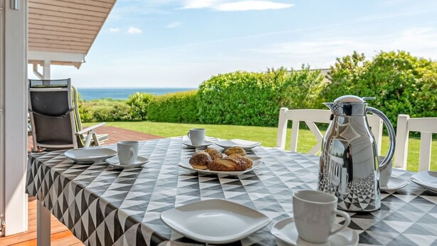 Une table dressée pour le petit-déjeuner, avec vue sur la mer, une cafetière en métal et une chaise.