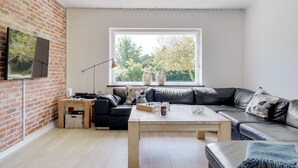 A modern living room with a brick wall, a large window, a black leather sofa, a wooden coffee table, and a TV mounted on the wall.