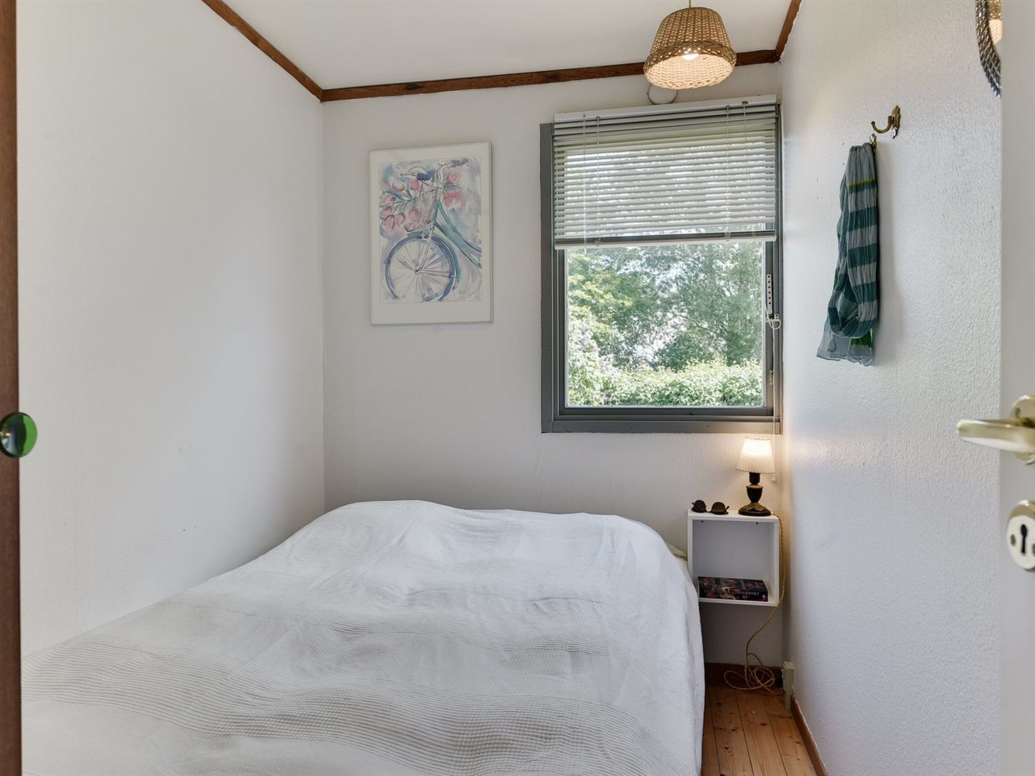 A bedroom with a bed, a window with blinds, a wall-mounted shelf with books, and a hanging towel.