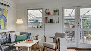 A modern living room with a sofa, armchair, and coffee table. There are bookshelves with books and decorative items, and a view of the outside through large windows.