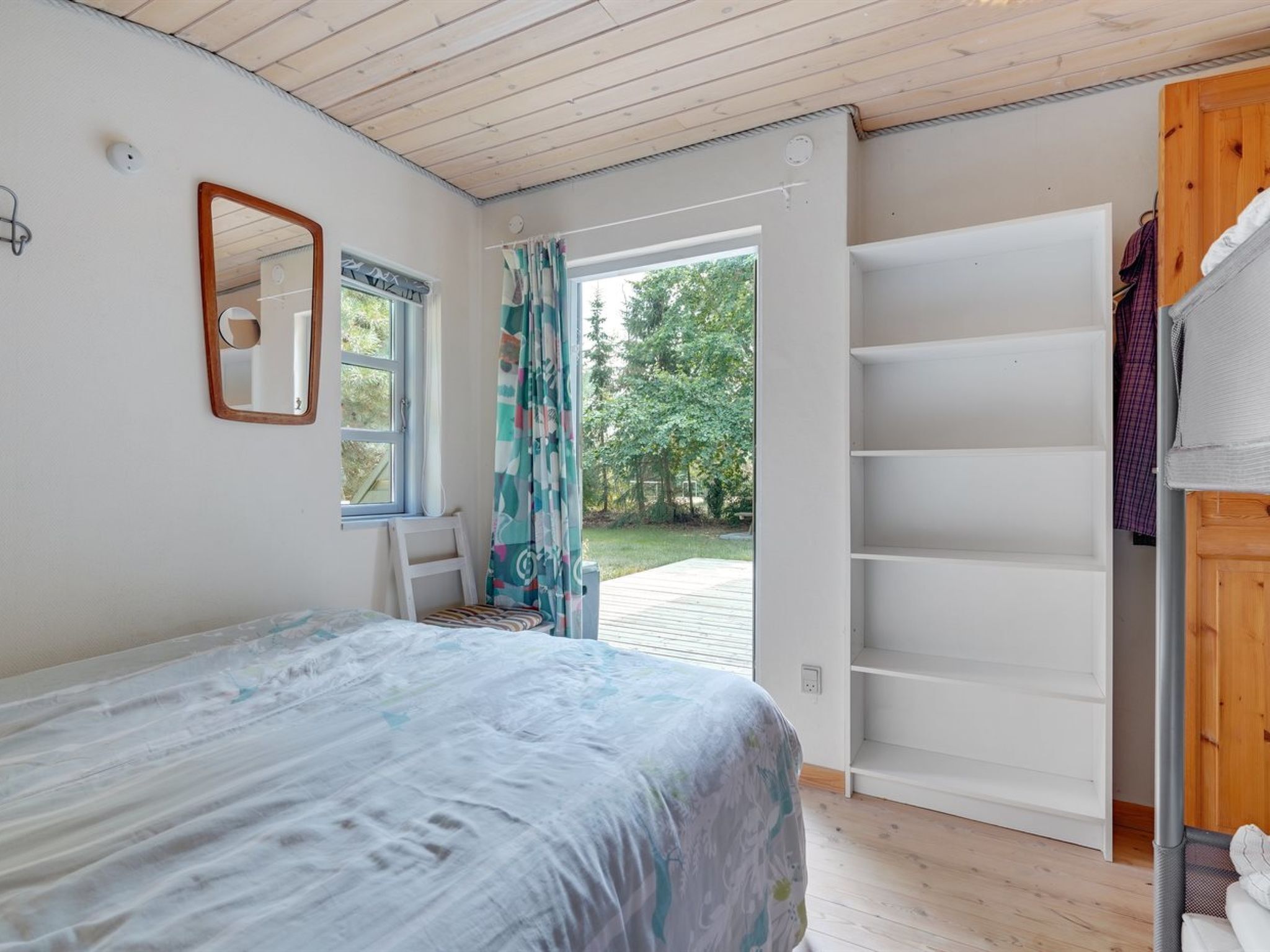A bedroom with a bed, a window with curtains, a mirror, and a wooden door leading outside.