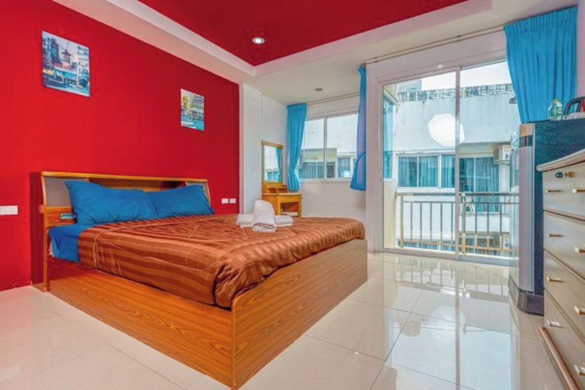 A hotel room with a wooden bed, a red accent wall, a balcony with blue curtains, and a view of the city.