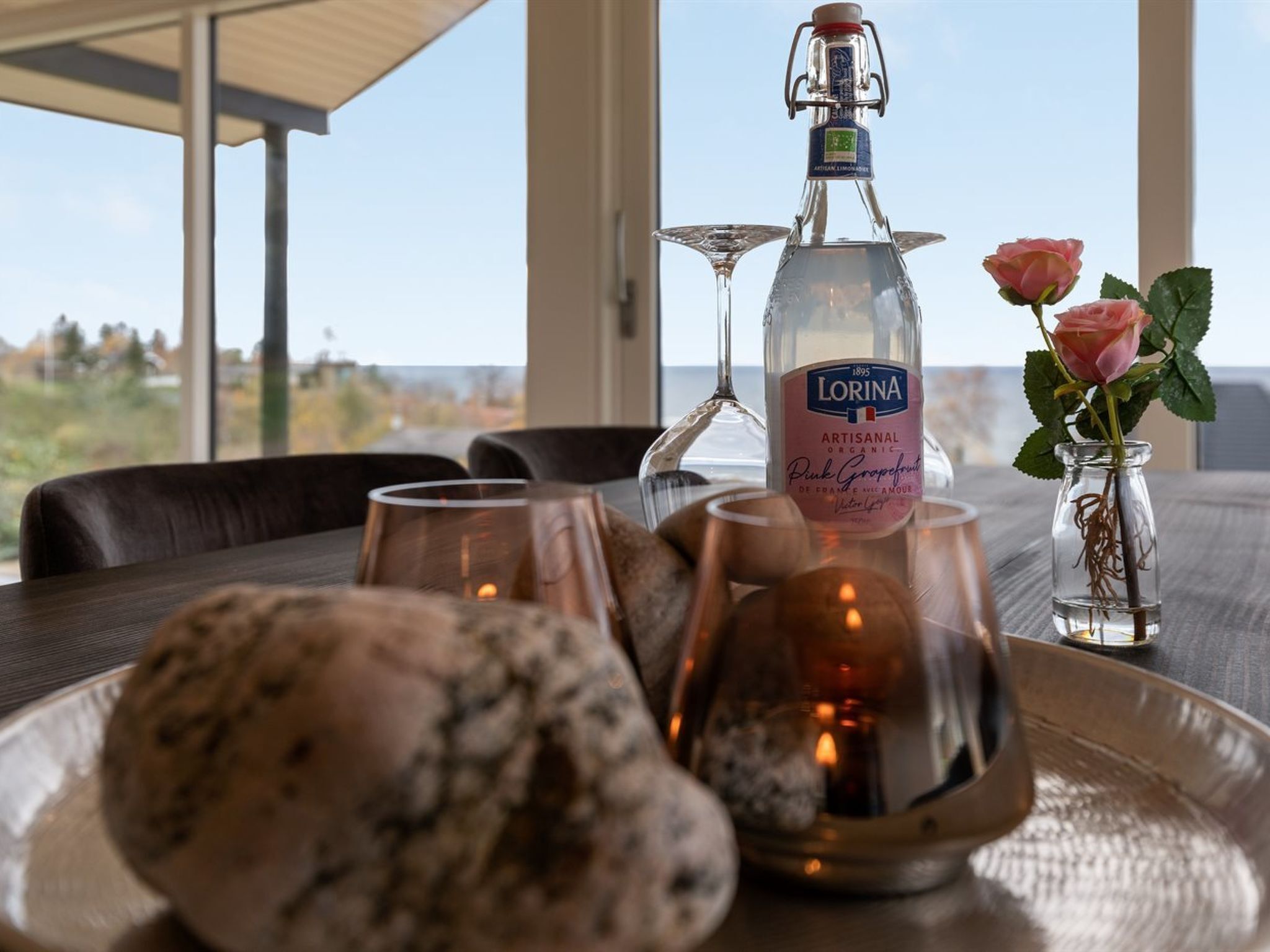 A table with a bottle of Lorina Pink Grapefruit, a glass, a candle, and a vase with roses.
