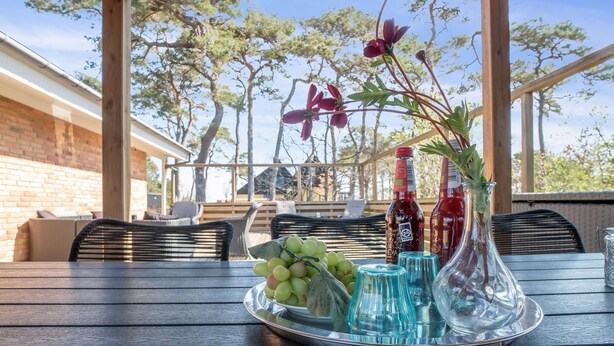 Una mesa con una bandeja de fruta, dos botellas y un florero con flores en un entorno al aire libre.
