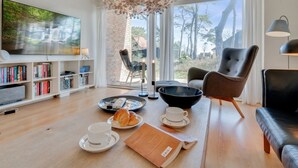 Una sala de estar moderna con un televisor grande, estanterías, una zona de comedor con cruasanes y café, y vistas a árboles a través de amplias ventanas.
