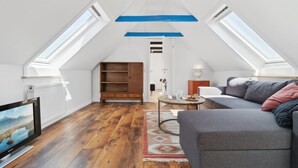 Un appartement moderne de style « loft » avec un plafond en pente, des fenêtres de toit, un plancher en bois, un canapé gris, une petite table et une bibliothèque.