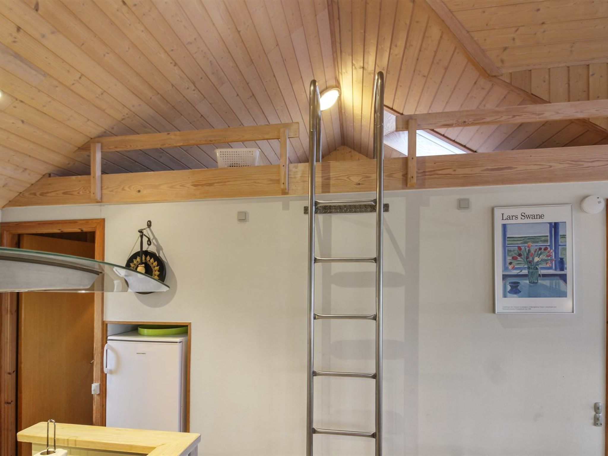 Een kamer met een houten plafond, een witte muur, een ladder, een koelkast, een wastafel en een poster.