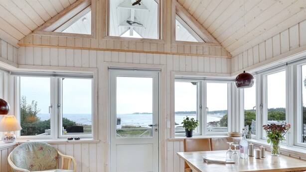 Une pièce chaleureuse avec un plafond en bois, de grandes fenêtres donnant sur la mer, une table à manger avec des chaises et une porte menant à l’extérieur.