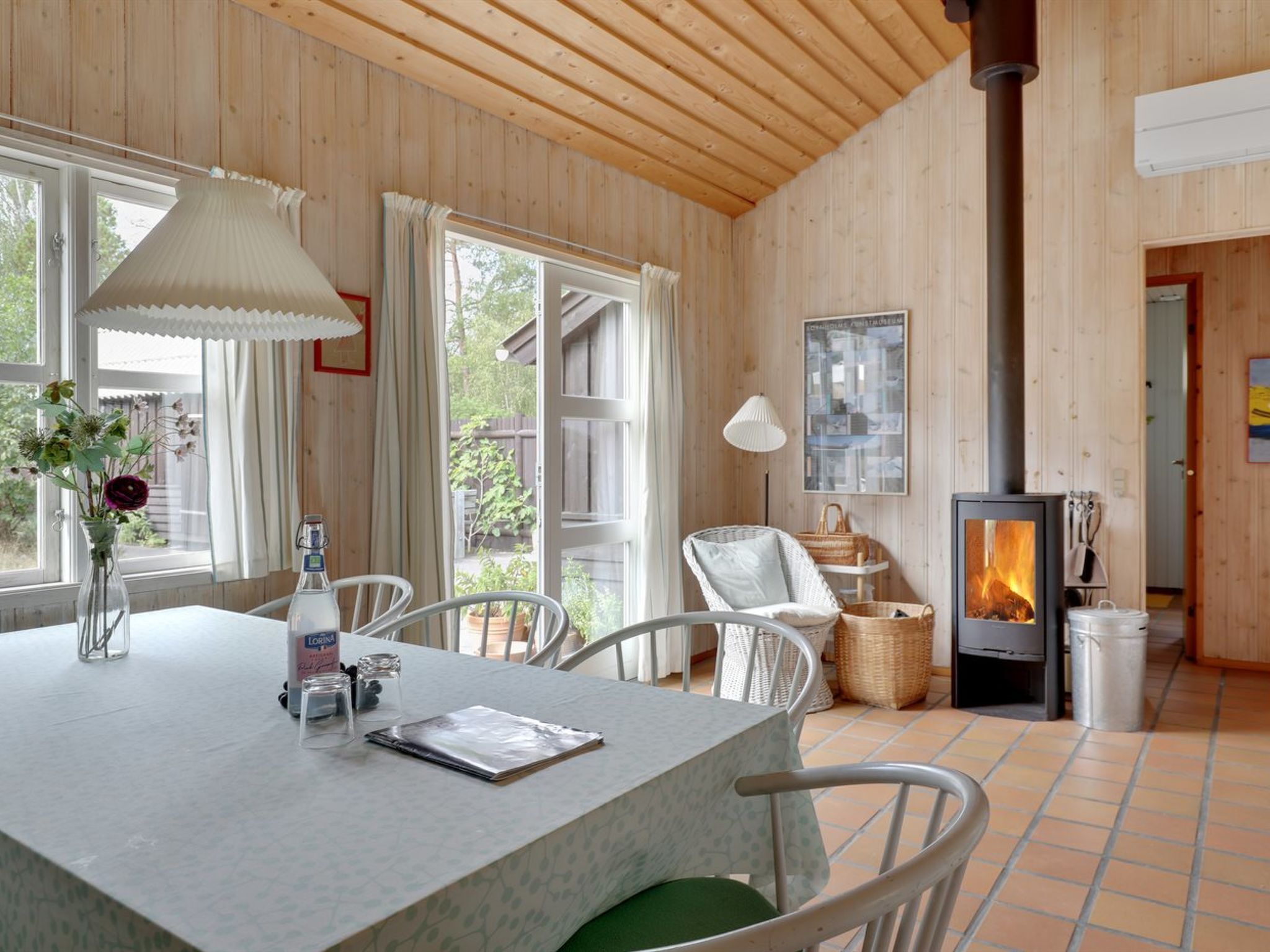 A cozy room with a wooden interior, a table with a vase of flowers, a chair, a table lamp, and a wood-burning stove.