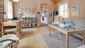 L’intérieur d’un chalet accueillant, avec un plafond en bois, un coin repas comprenant une table et des chaises, un canapé avec des coussins, une bibliothèque et, au fond, une cuisine.