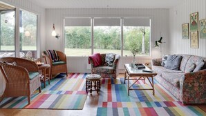 Un salon chaleureux, meublé de meubles en osier, agrémenté d’un tapis coloré et doté de grandes fenêtres offrant une vue sur la verdure.